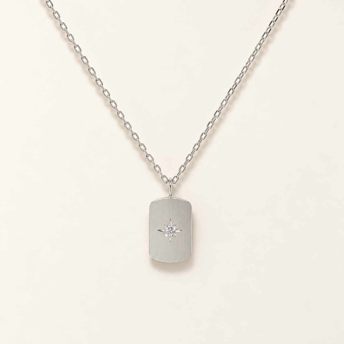 A silver dog tag necklace with a small starburst diamond accent, displayed on a clean cream background.