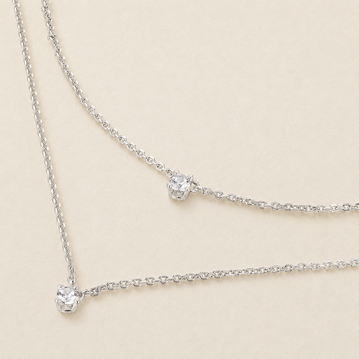 A delicate silver-toned double-layer necklace with two small round clear gemstones, displayed on a light beige background.
