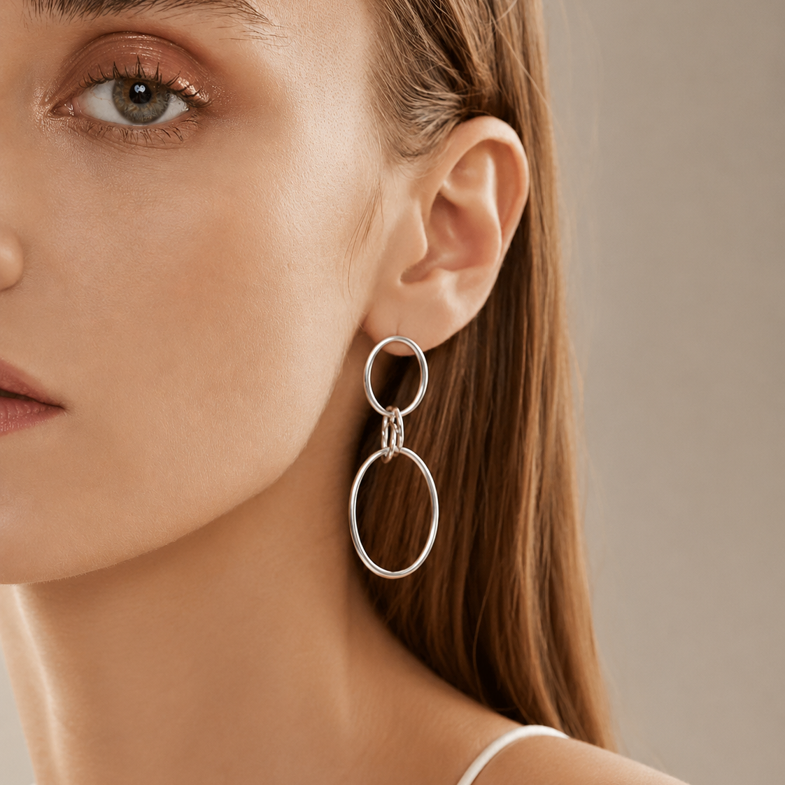 A close-up of a woman wearing a large silver-toned dangle earring with two interlocking oval rings, shown against a soft neutral background.