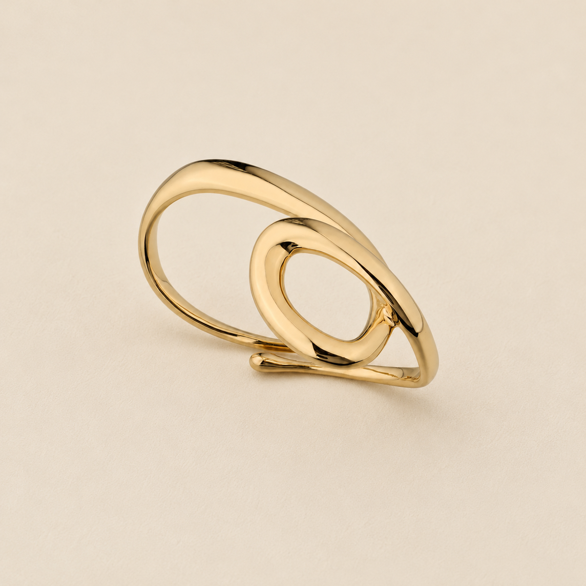 A polished gold-toned ring with an abstract, intertwined looped design, photographed on a clean, light beige textured background.