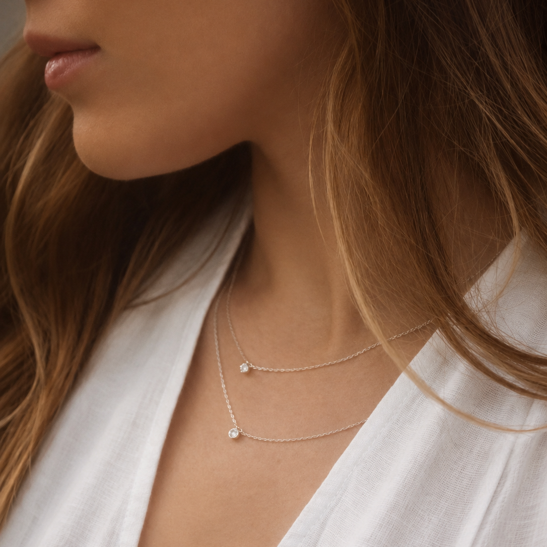 A close-up lifestyle shot of a woman wearing a delicate double-layered silver necklace with small clear gemstone pendants, styled with a white linen top.