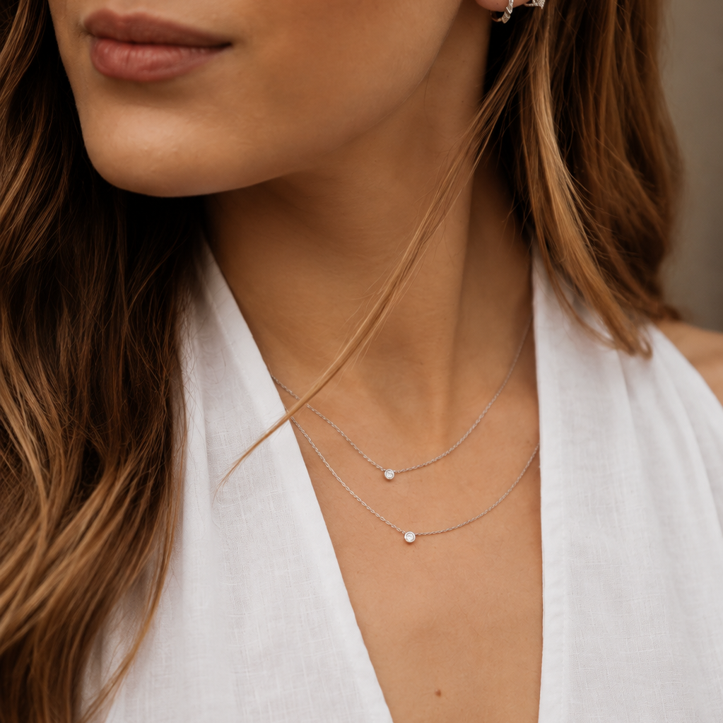 A close-up of a woman wearing delicate silver layered necklaces with small pearl-like beads, along with small hoop earrings, styled with a white halter top.