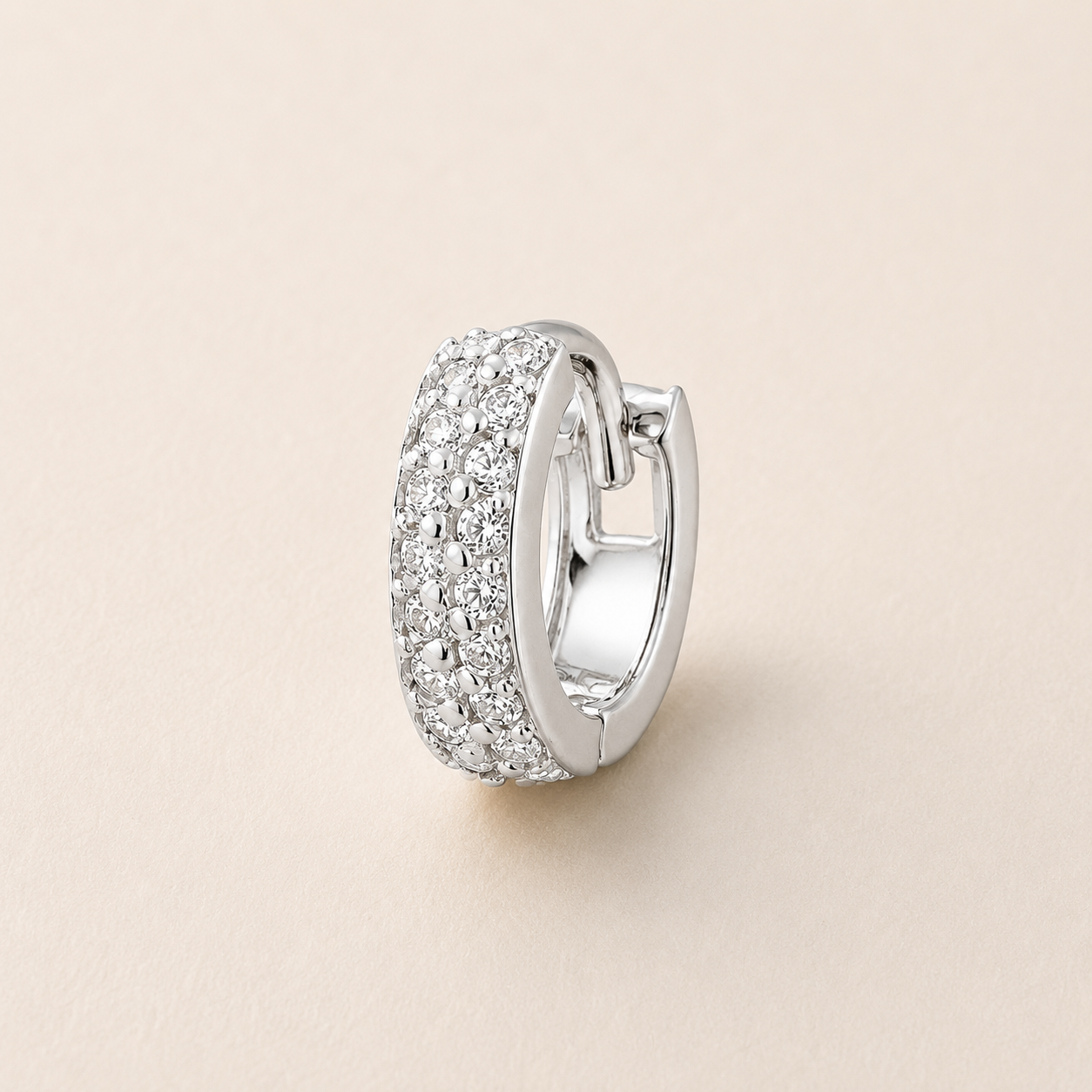 A silver huggie hoop earring paved with clear cubic zirconia stones, shown on a light beige background.