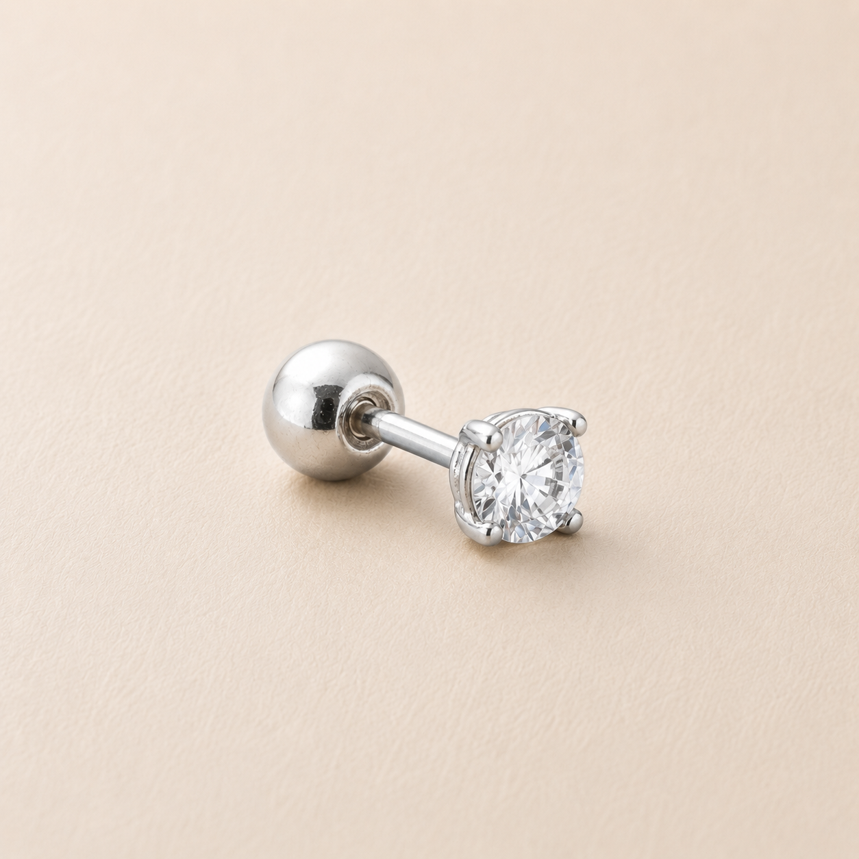A single silver-toned labret stud earring with a round brilliant-cut clear gemstone in a four-prong setting, shown on a warm beige textured background.