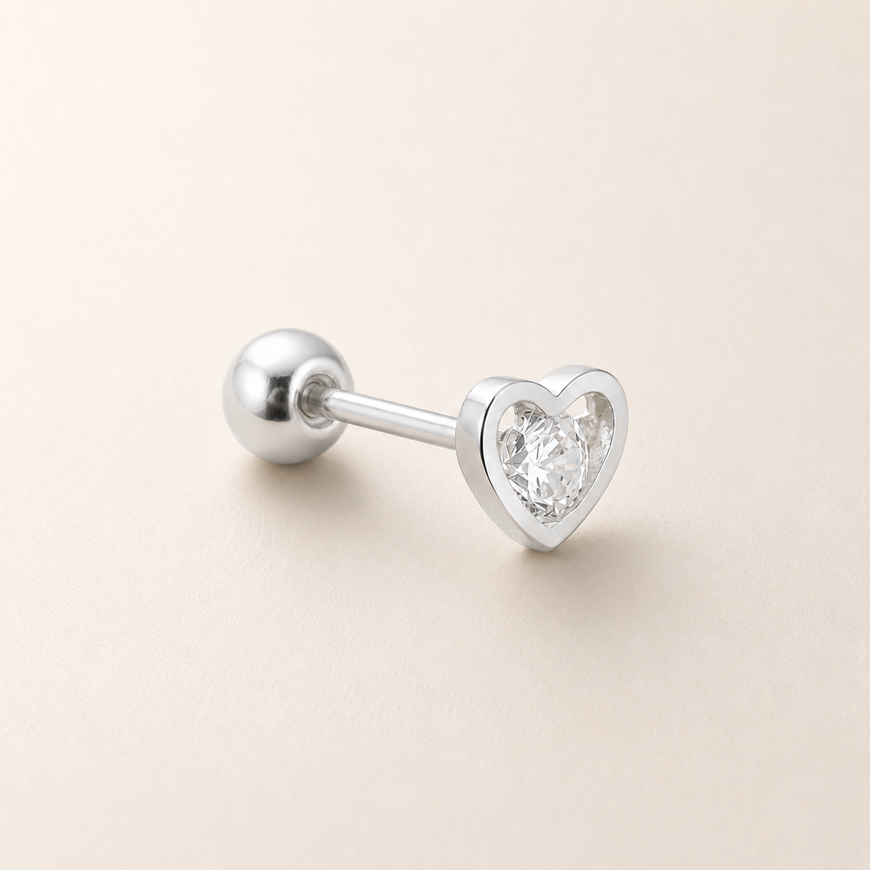 A silver-toned labret stud earring with a heart-shaped clear gemstone in a bezel setting, shown on a light beige background.