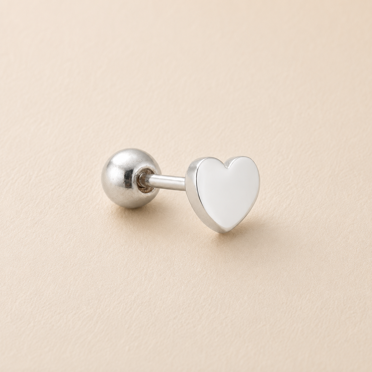 A silver-toned flat-back labret stud earring featuring a polished heart-shaped face and a spherical ball back, displayed on a textured beige surface.