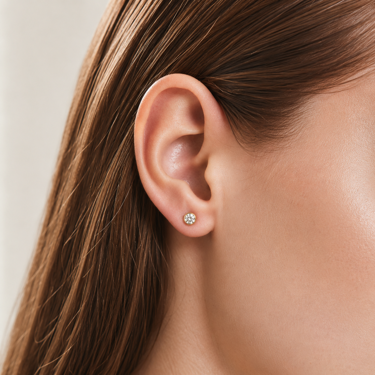 A close-up of a person's ear wearing a small round stud earring with a sparkling clear gemstone in a silver-toned setting.