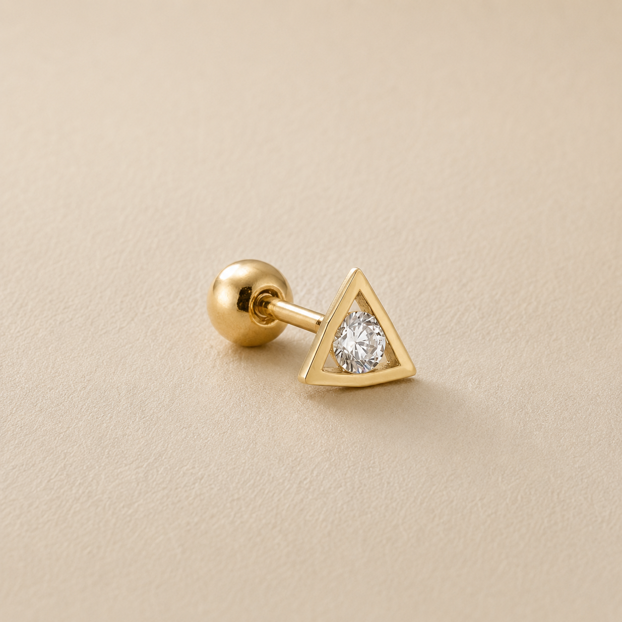 A gold stud earring featuring a small triangular bezel-set clear gemstone on a polished post with a round ball back, displayed on a textured beige surface.