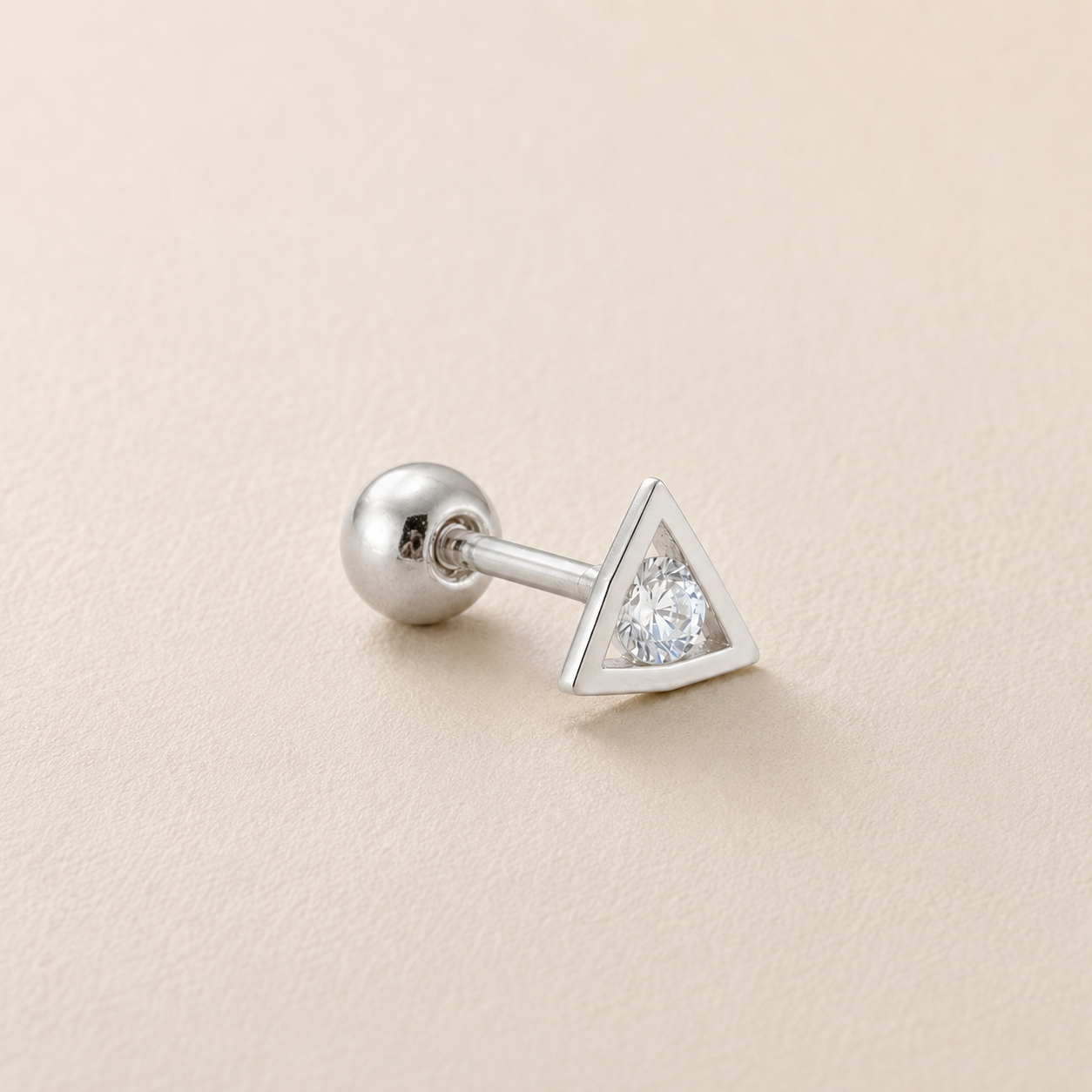 A silver-toned triangular stud earring with a clear gemstone, shown on a light beige textured background.