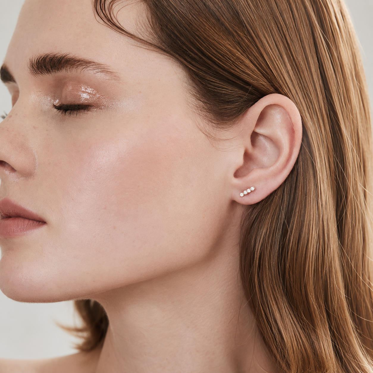 Close-up of a woman's ear wearing a small silver stud earring with three tiny clear stones, shown against soft neutral background.