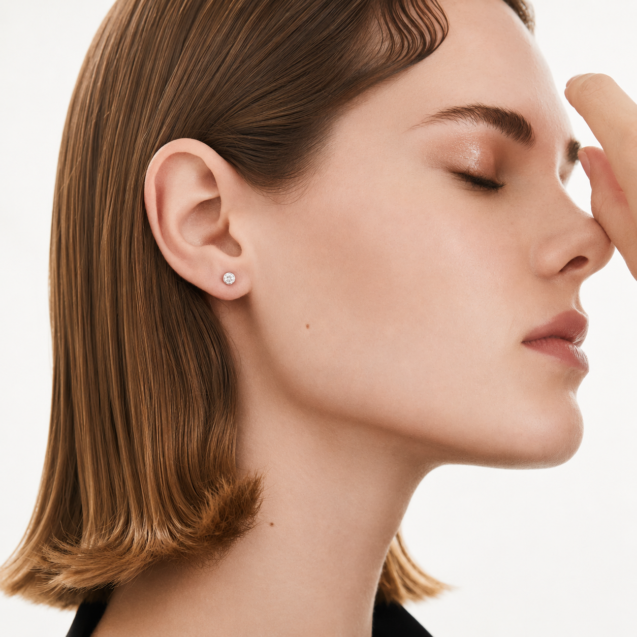 A close-up side profile of a woman with brown hair wearing a small round silver stud earring, with her hand near her face against a clean white background.