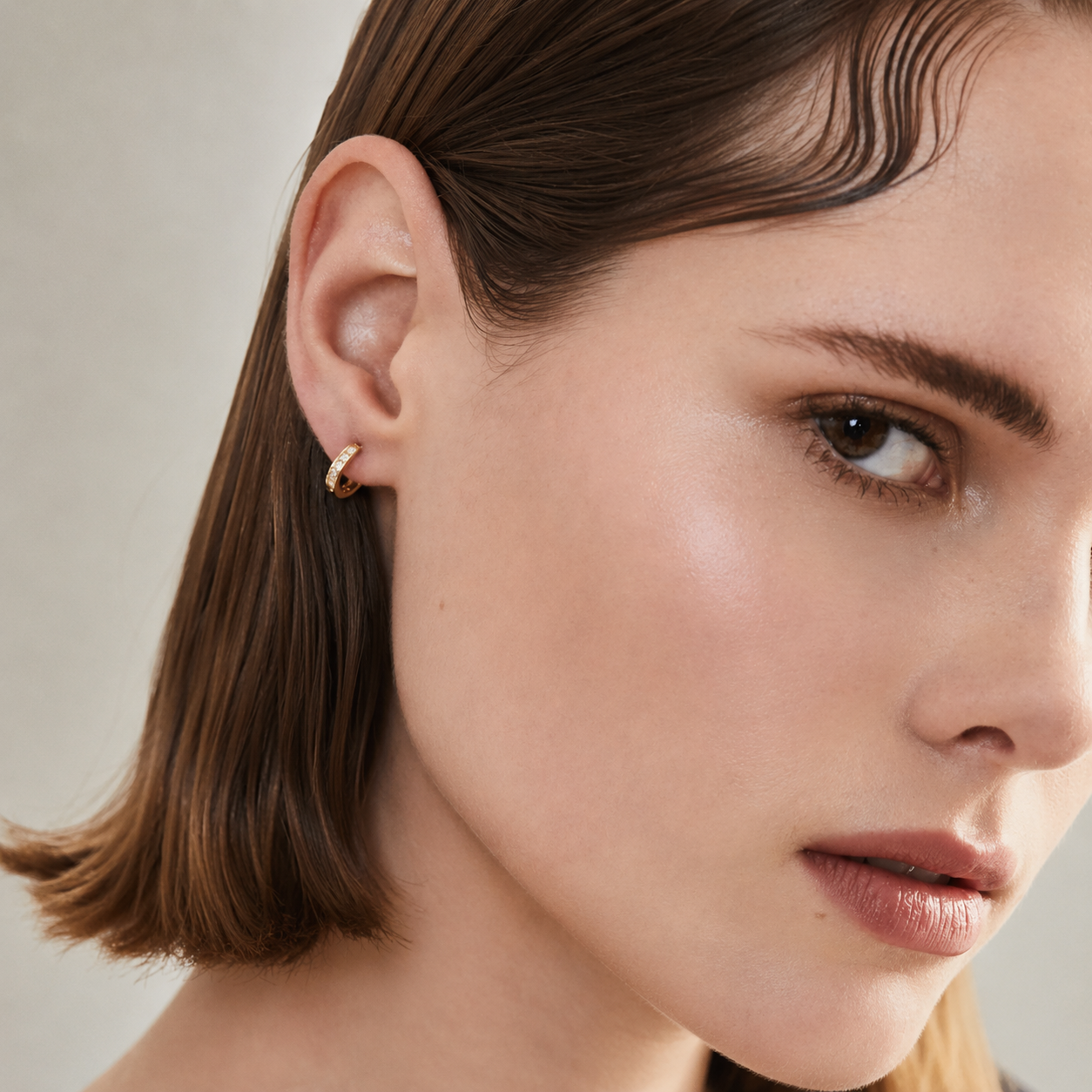A close-up of a woman's ear wearing a small gold huggie hoop earring, with her hair styled in a side part and a soft neutral background.