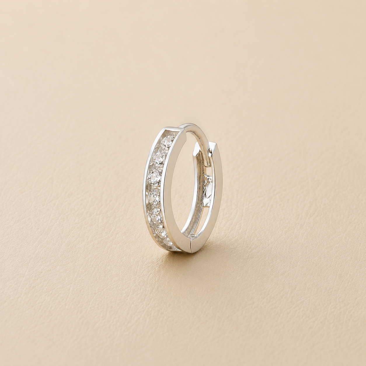 A silver huggie hoop earring with a channel-set row of clear cubic zirconia stones, displayed on a textured beige surface.