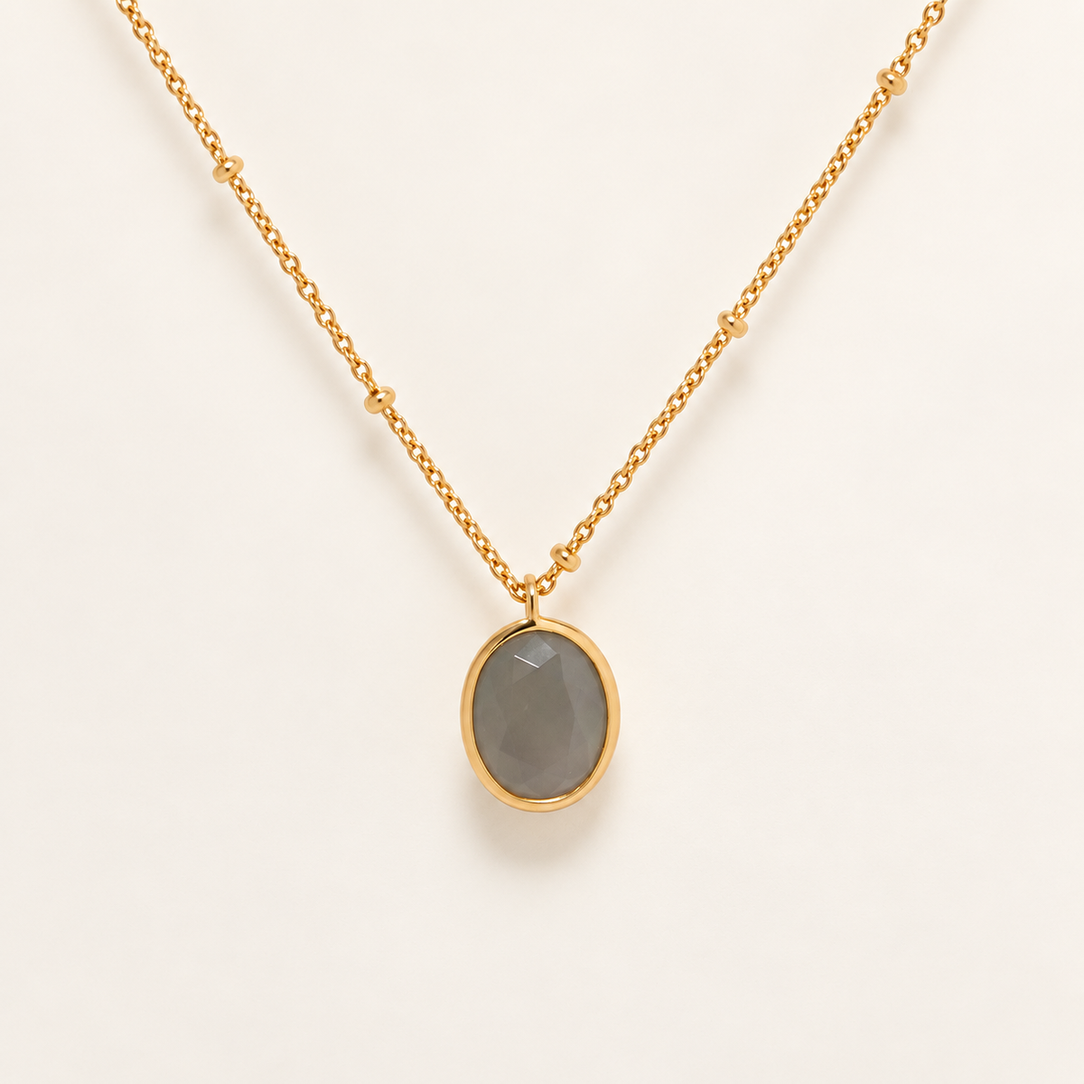 A gold chain necklace with a bezel-set oval gray stone pendant, displayed against a clean off-white background.