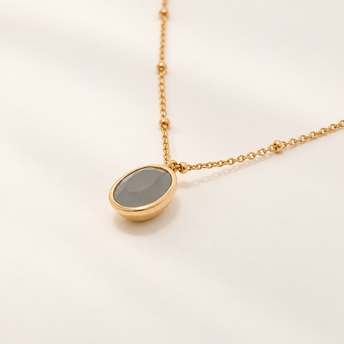 A gold chain necklace featuring an oval pendant with a smooth, matte gray stone in a polished gold bezel, set against a clean, off-white background.