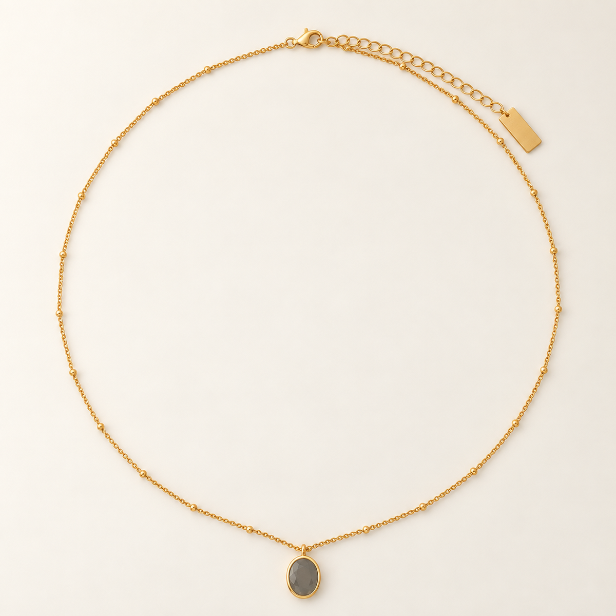 A delicate gold chain necklace with a small oval gray stone pendant, displayed on a clean off-white background.