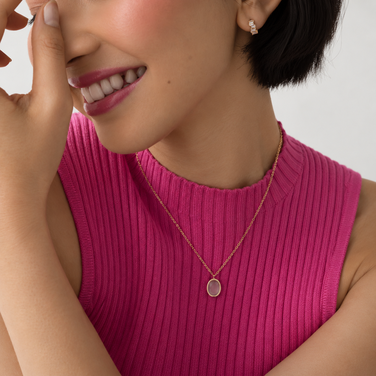 A close-up lifestyle shot of a woman wearing a delicate rose gold chain necklace with a flat oval pendant, paired with a bright pink ribbed knit top.