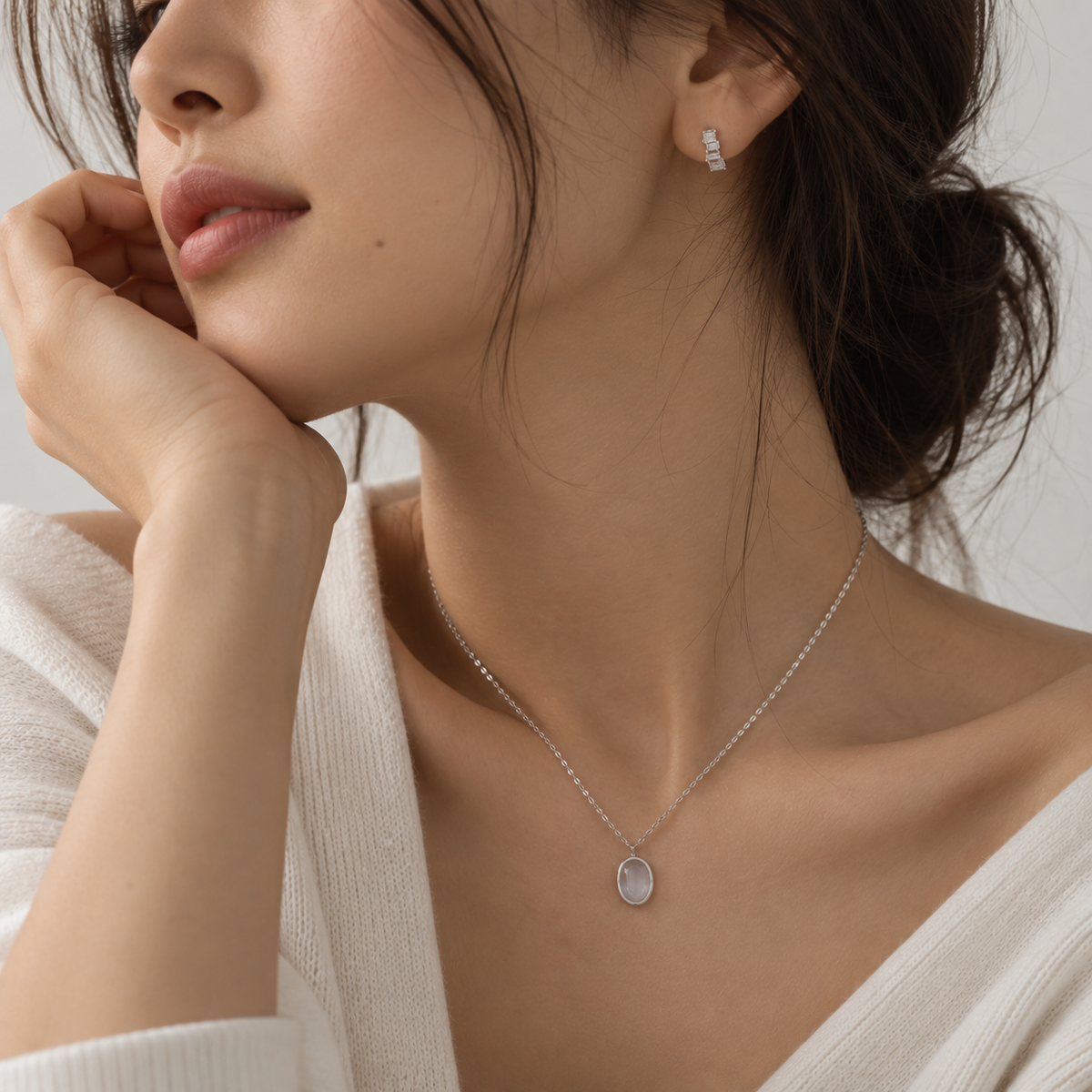 A close-up lifestyle shot of a woman wearing a delicate silver necklace with a small oval pendant and a matching silver stud earring, styled with a white knit top against a soft neutral background.
