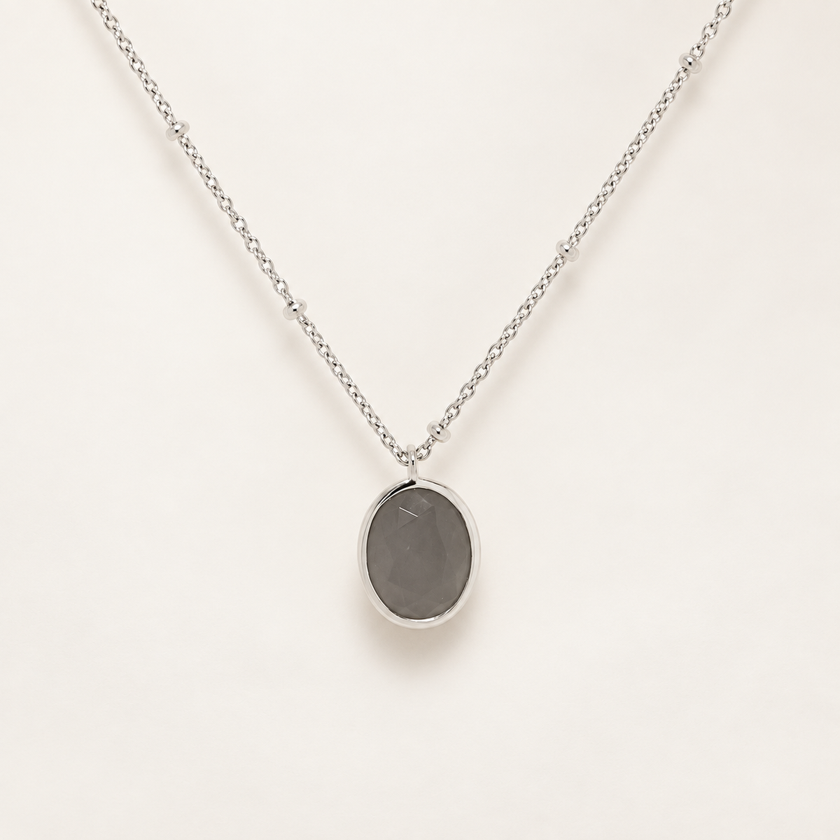 A silver chain necklace with an oval pendant featuring a dark gray stone in a silver bezel, displayed on a clean off-white background.