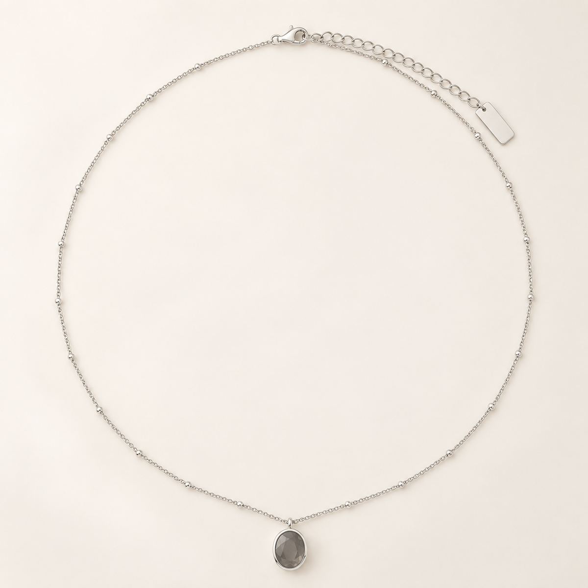 A delicate silver chain necklace with a small oval gray stone pendant, displayed on a clean, light beige background.