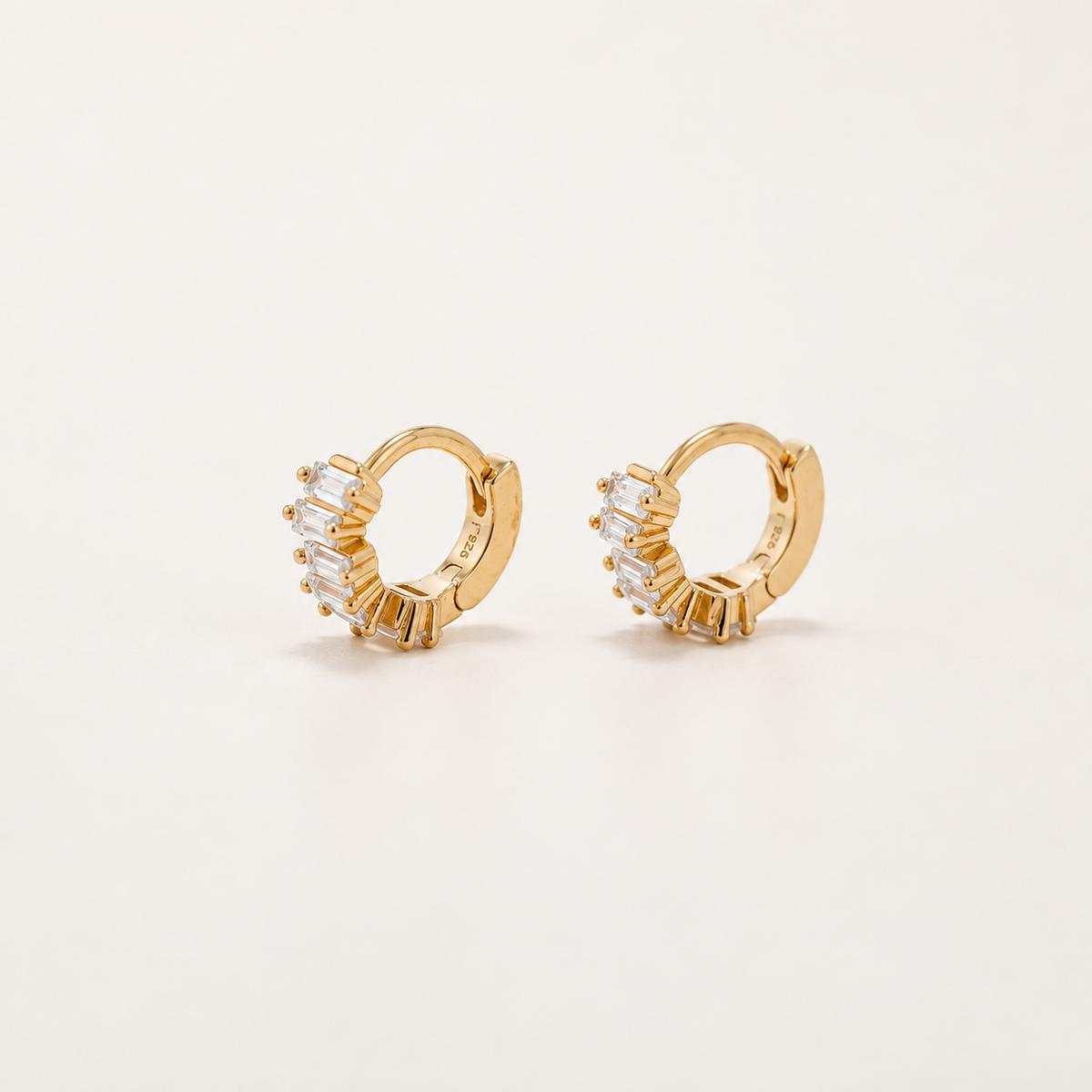 A pair of gold huggie hoop earrings with baguette-cut clear crystals, displayed on a clean off-white background.