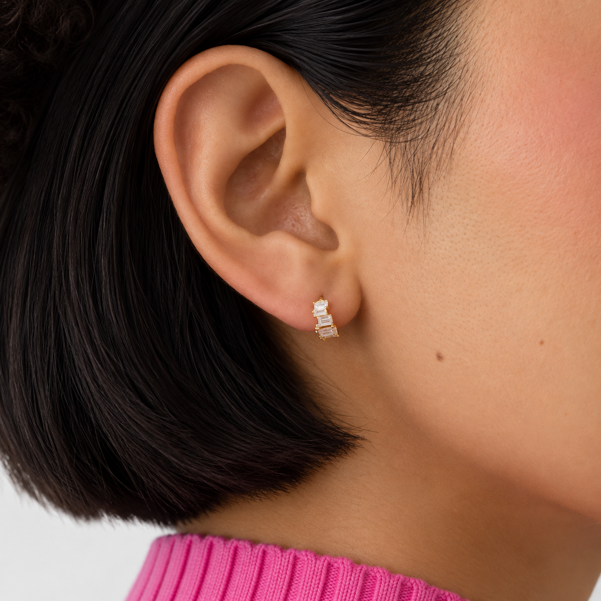 A close-up of a person's ear wearing a small gold huggie hoop earring with clear rectangular stones.