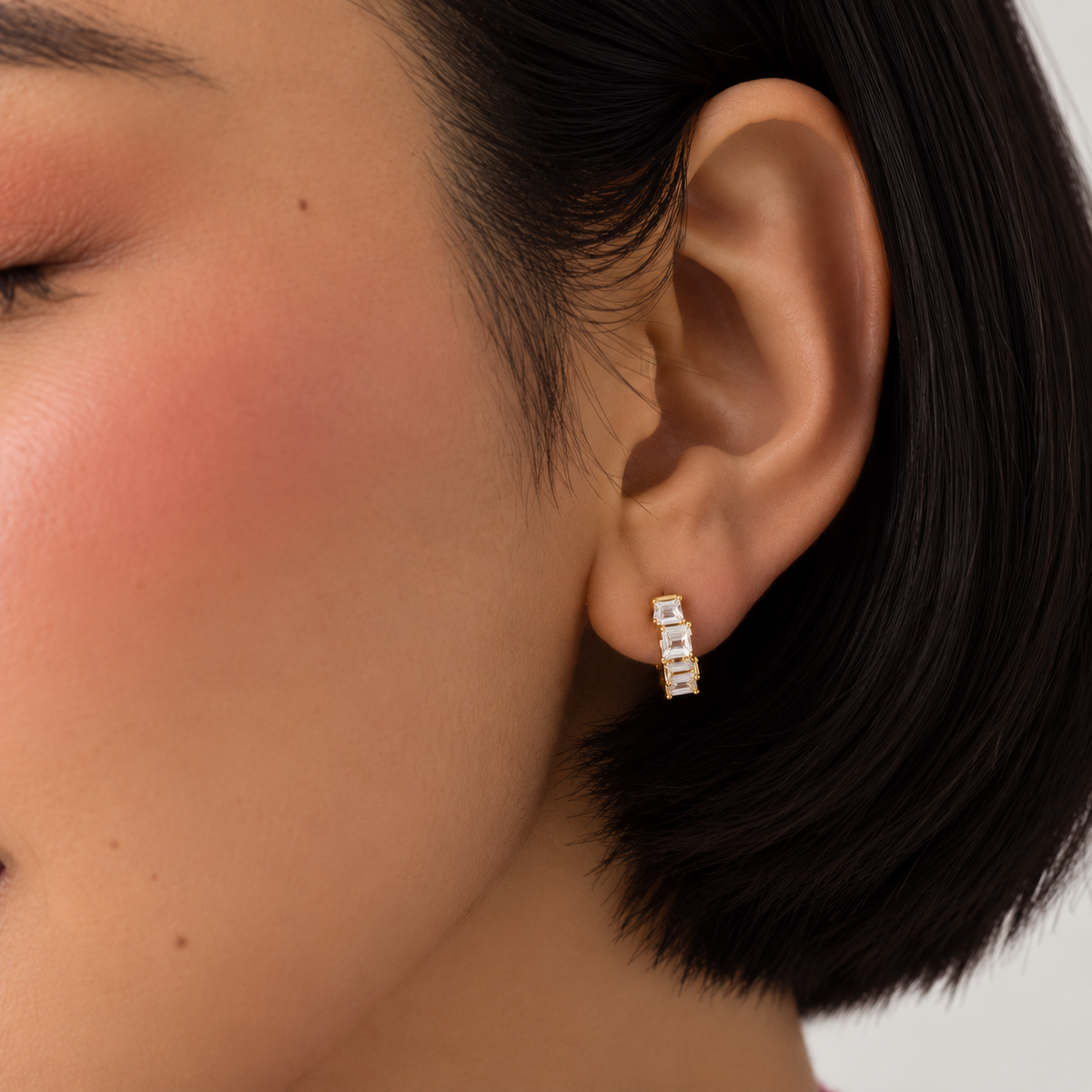 A close-up of a model's ear wearing a small gold huggie hoop earring set with clear baguette-cut stones.