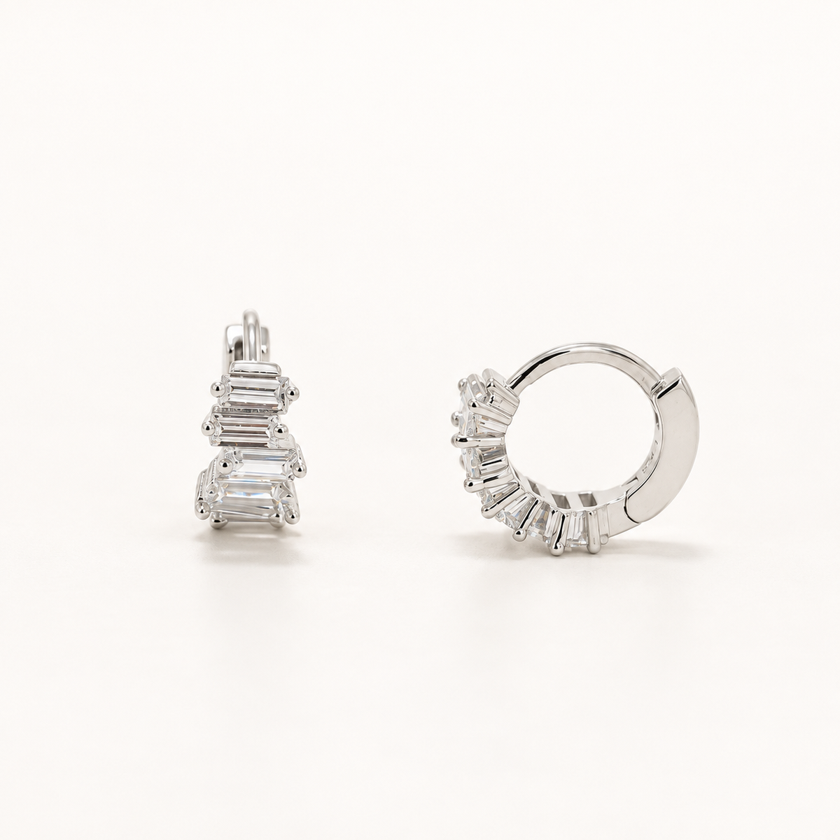 A pair of silver huggie hoop earrings with baguette-cut clear gemstones, shown from two angles on a clean white background.