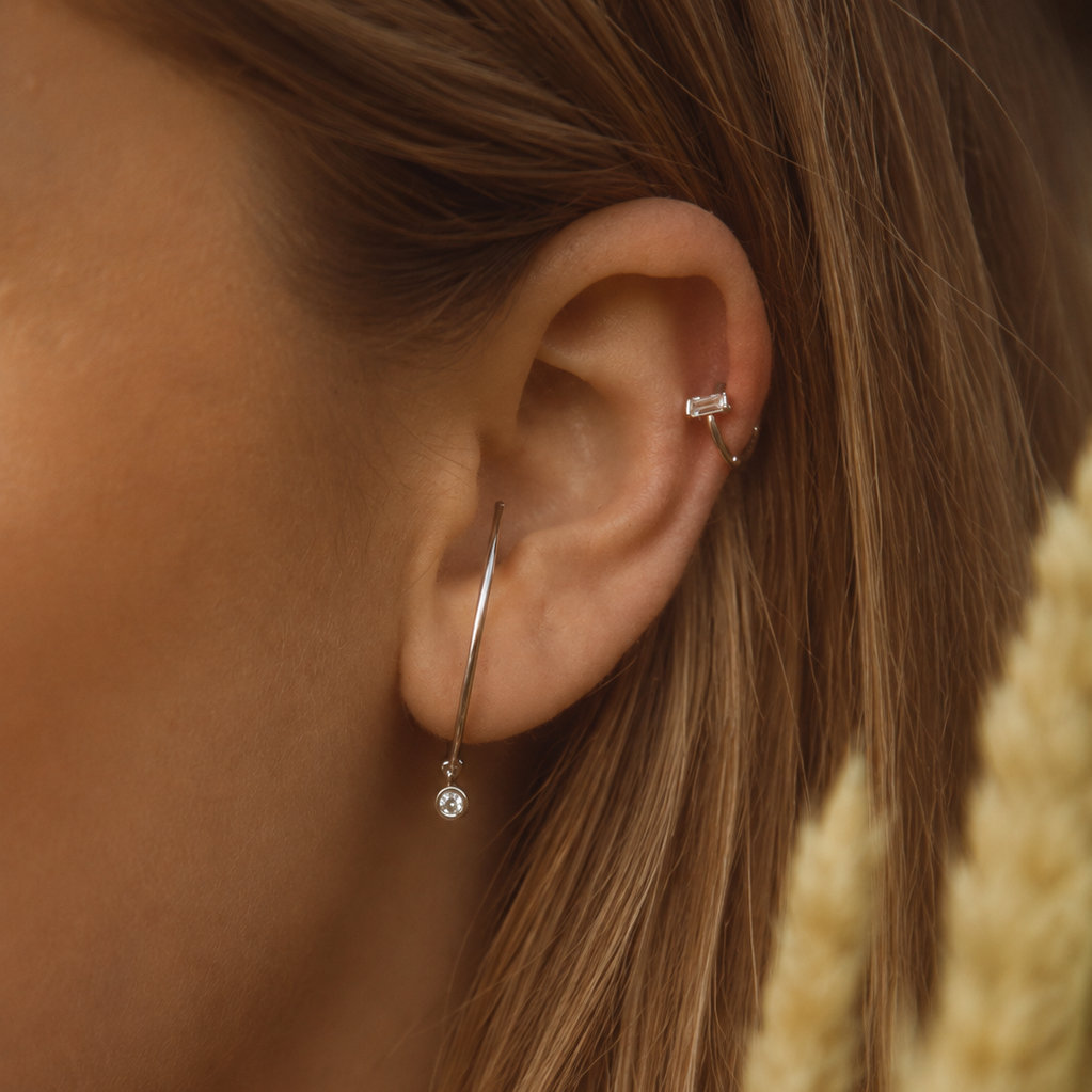 A close-up of a person's ear showcasing a minimalist silver hoop earring with a small clear crystal drop, worn alongside a silver cartilage piercing. The background is softly blurred with warm golden dried grasses.