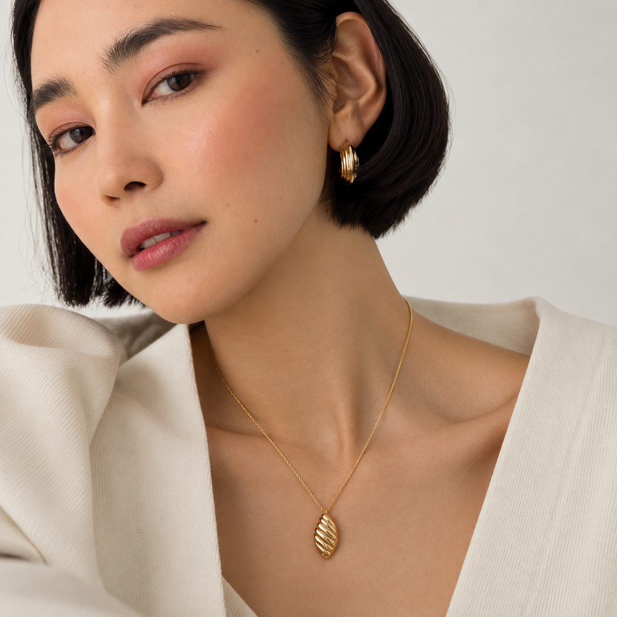 A close-up of a woman wearing a delicate gold necklace with a textured leaf-shaped pendant and a matching gold hoop earring, styled with a cream-colored top against a soft, light background.
