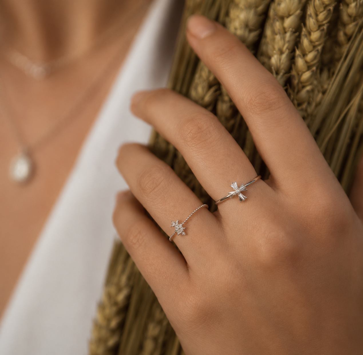 A close-up of a hand wearing two delicate silver rings with small floral or starburst motifs, posed against a background of dried wheat stalks.
