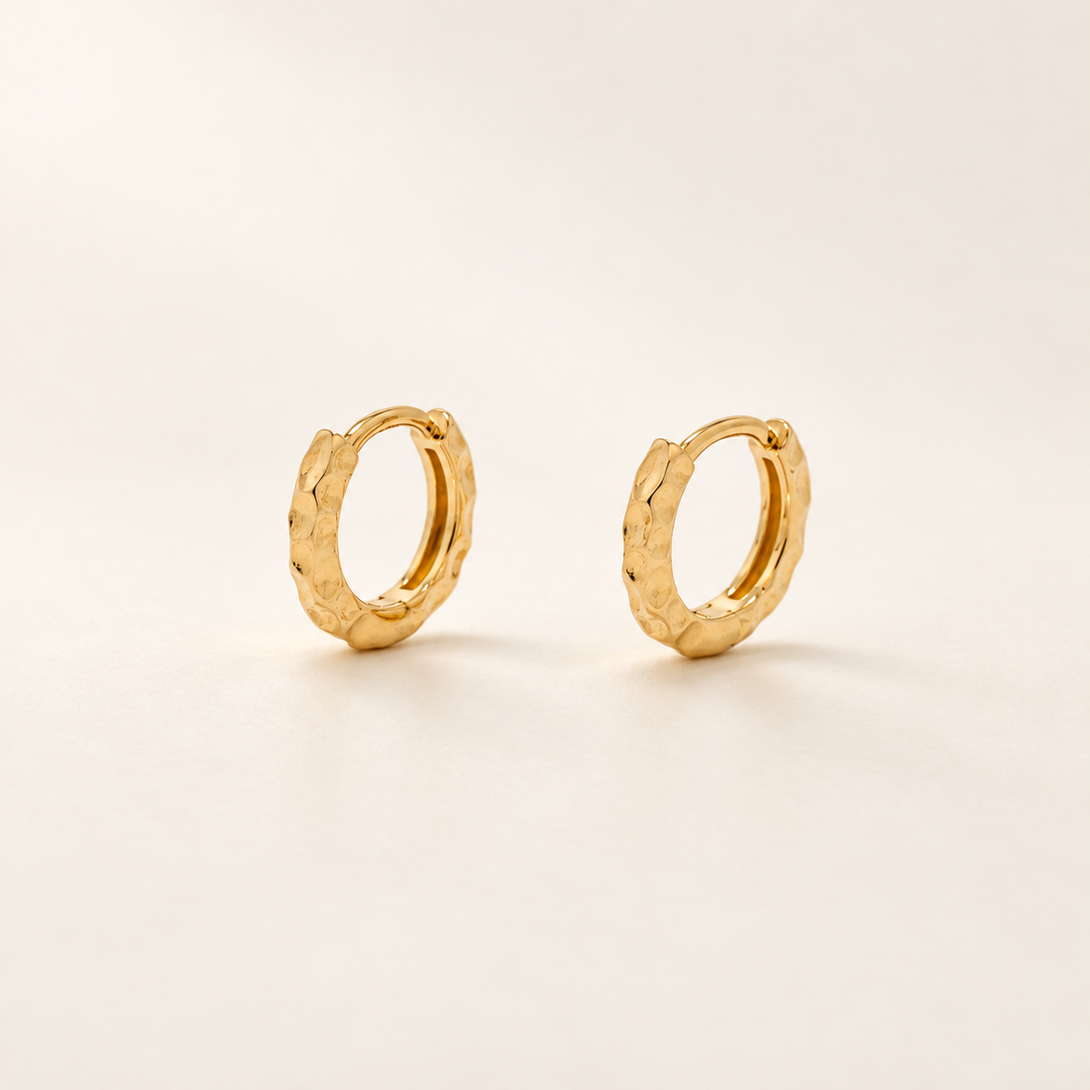 A pair of textured gold huggie hoop earrings on a clean, light beige background.