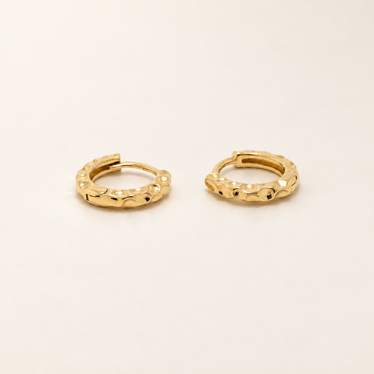 A pair of textured gold huggie hoop earrings on a clean, solid off-white background.