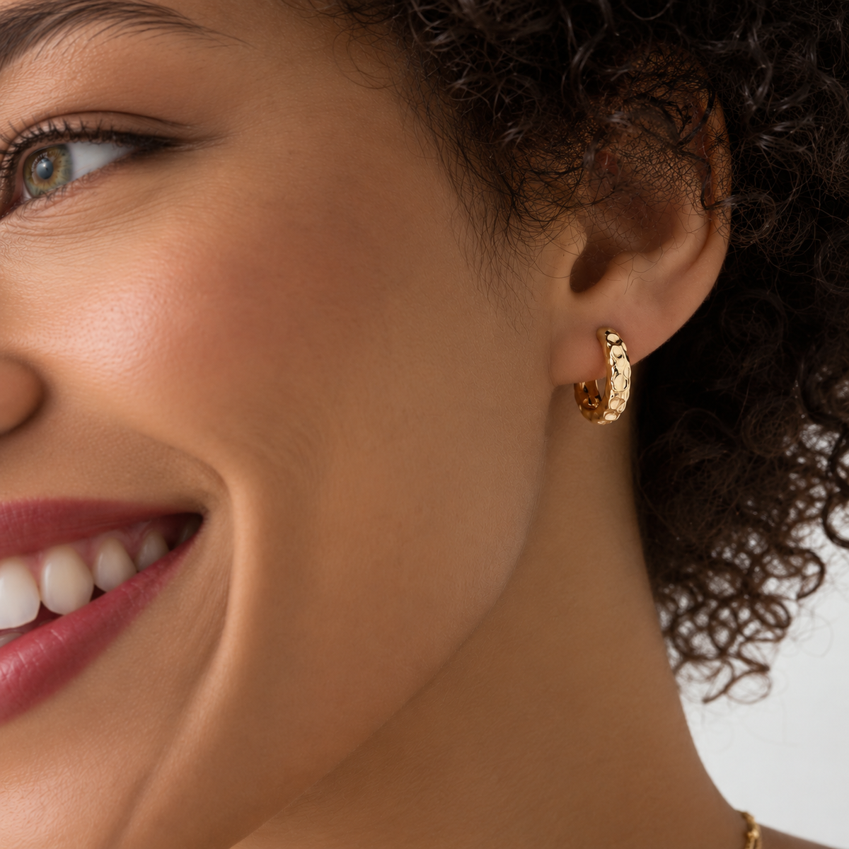 A close-up of a woman's ear wearing a small textured gold huggie hoop earring, with her face and curly hair partially visible against a light background.