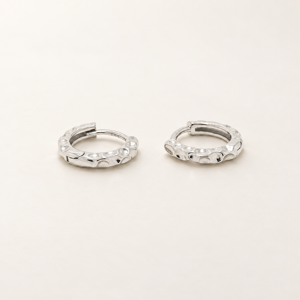 A pair of textured silver huggie hoop earrings on a clean, light beige background.