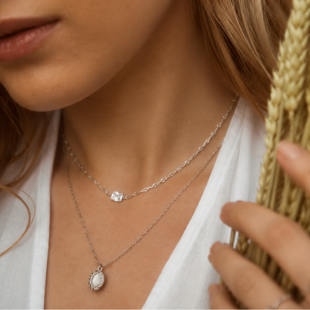 Close-up of a woman wearing layered silver necklaces, including a delicate chain with a small square-cut stone and a longer chain with a white oval pendant, along with a small hoop earring, holding dried wheat stalks.