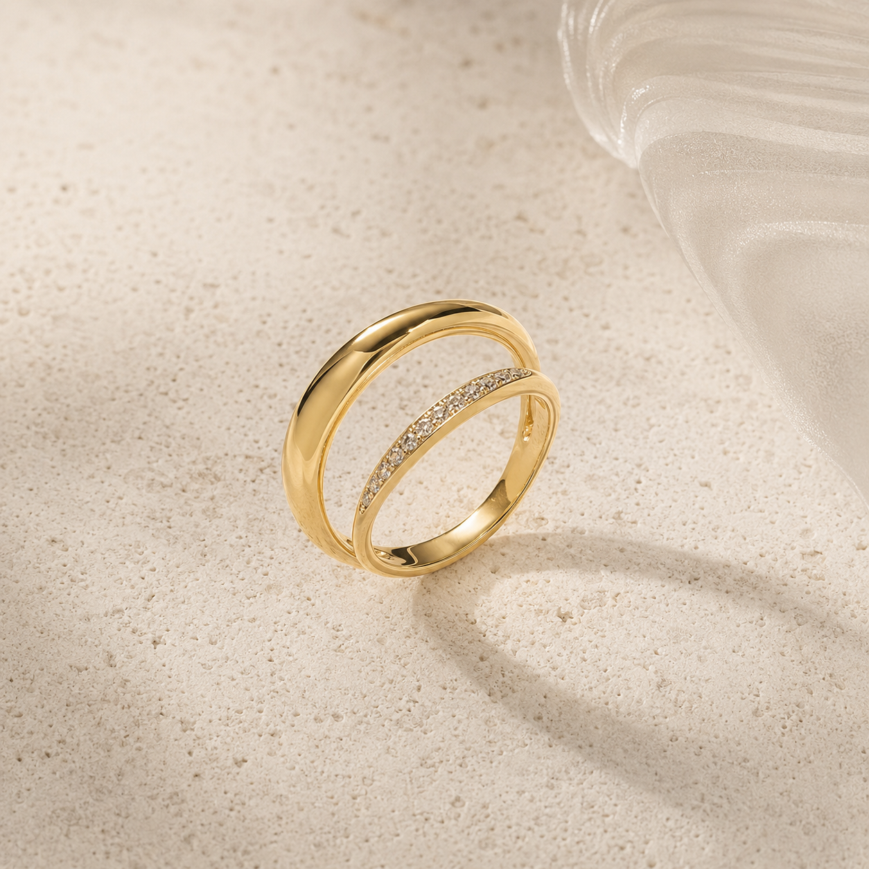 A gold double-band ring with a pavé-set row of small clear stones on one band, photographed on a light beige textured surface with a soft shadow and part of a translucent glass object in the background.