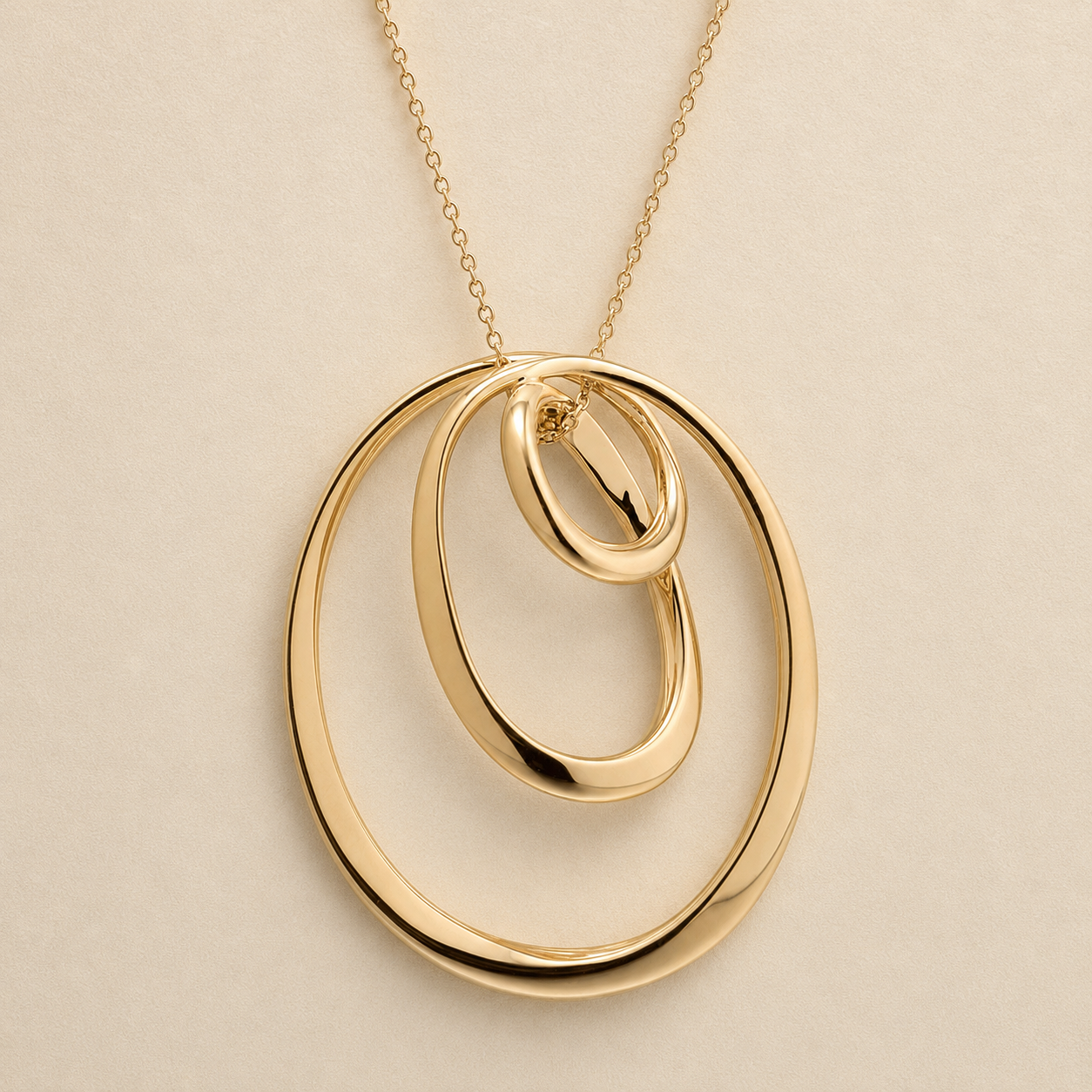 A gold pendant necklace featuring three interlocking, overlapping oval rings on a delicate chain, set against a solid light beige background.
