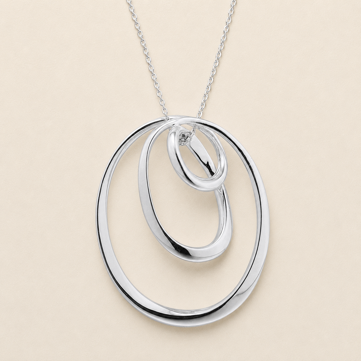 A silver pendant necklace featuring three interlocking, nested oval rings on a delicate silver chain, set against a solid light beige background.