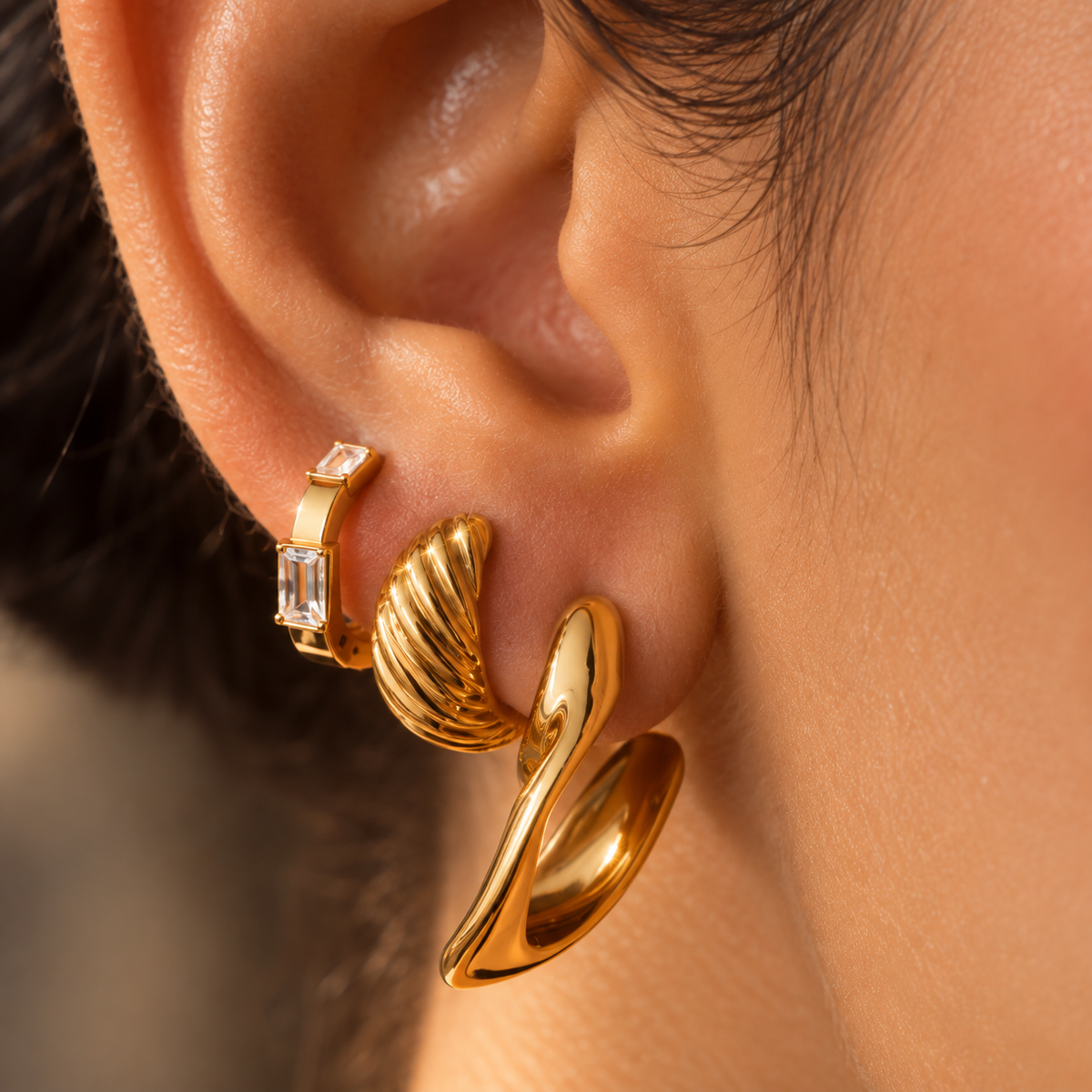 A close-up of a person's ear showcasing a stack of gold earrings, including a textured spiral hoop, a simple curved hoop, and a small gold huggie with a clear baguette gemstone.