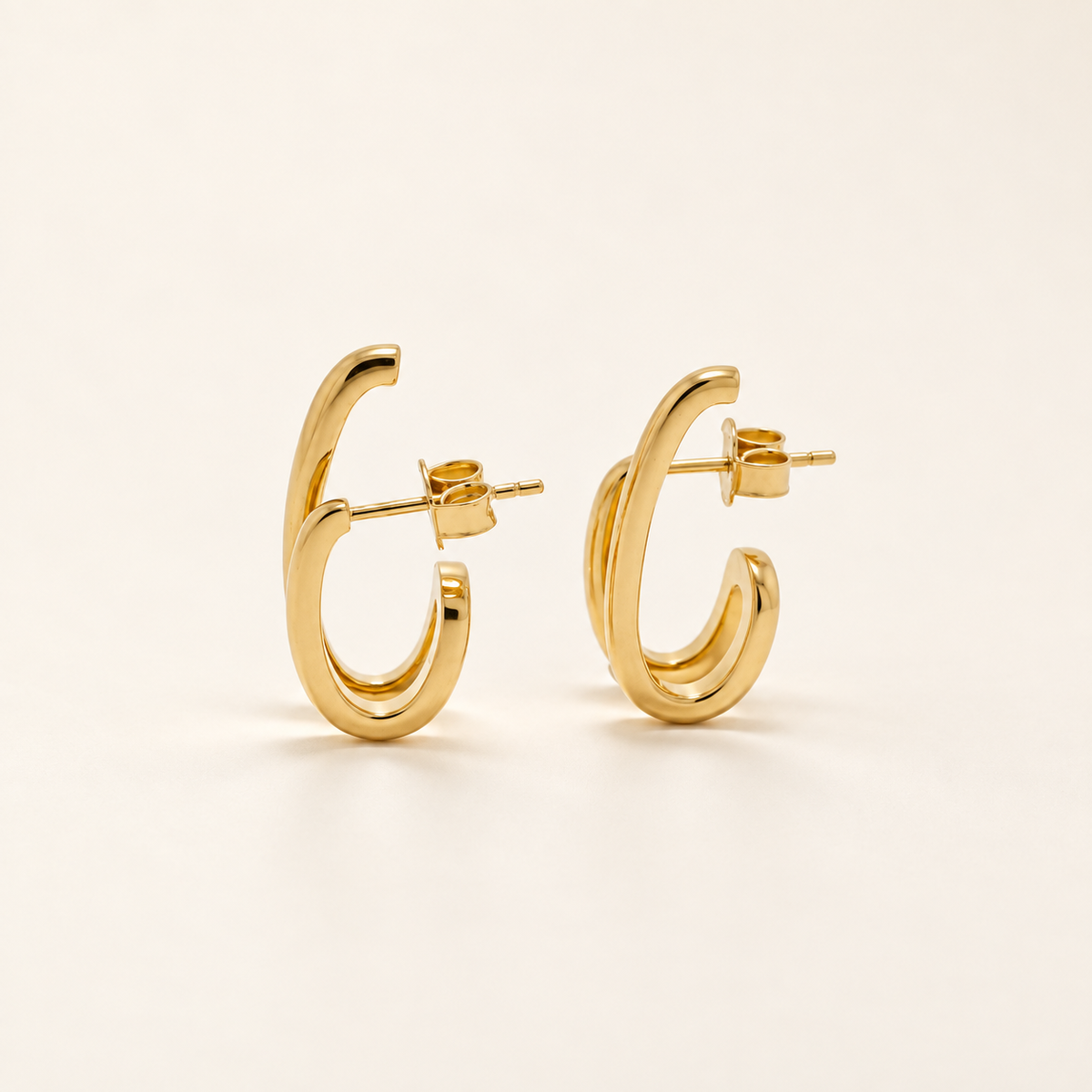A pair of polished gold hoop earrings with a thick, rounded double-band design, shown from a side profile on a clean, light beige background.