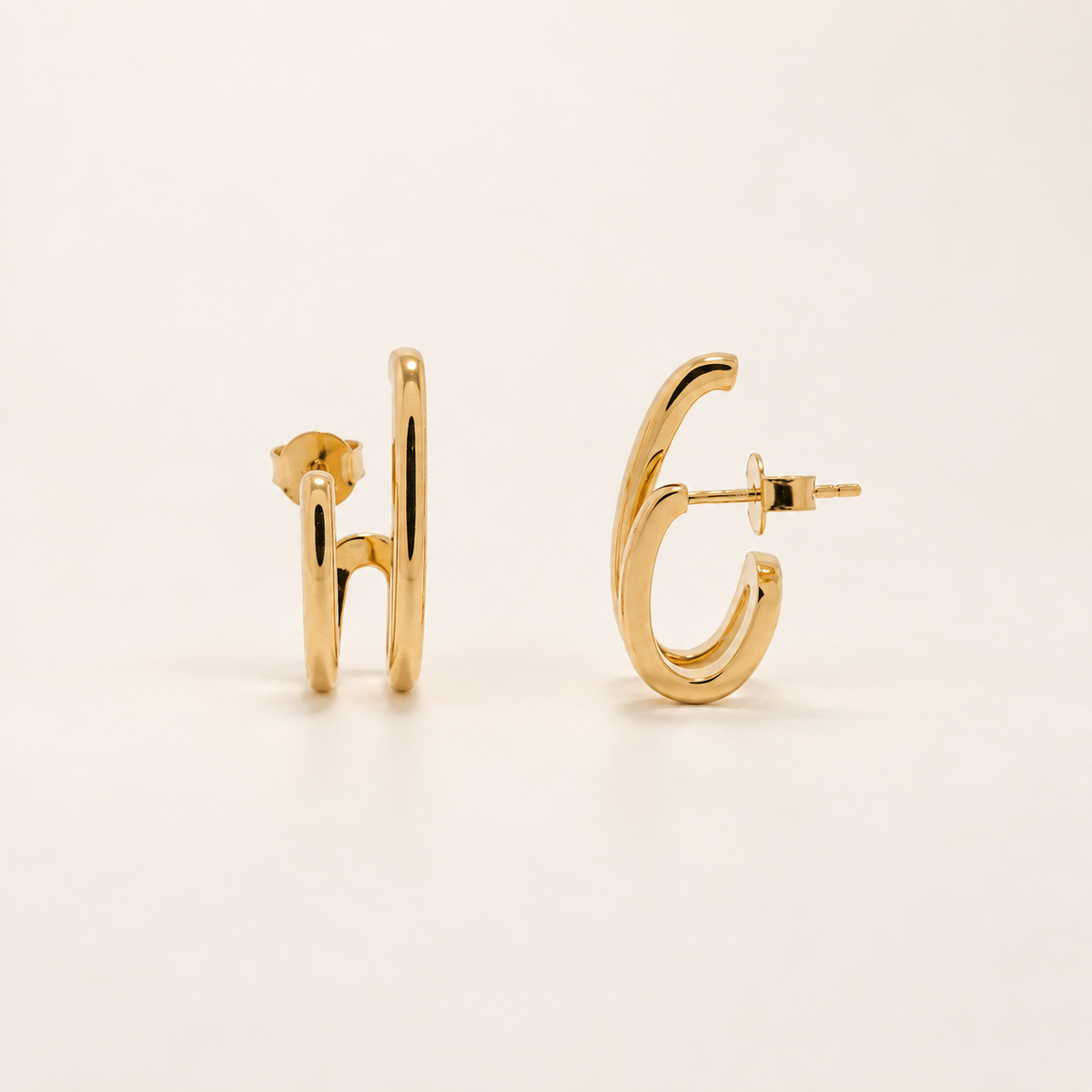 A pair of gold huggie hoop earrings shown from the side on a clean, light beige background.