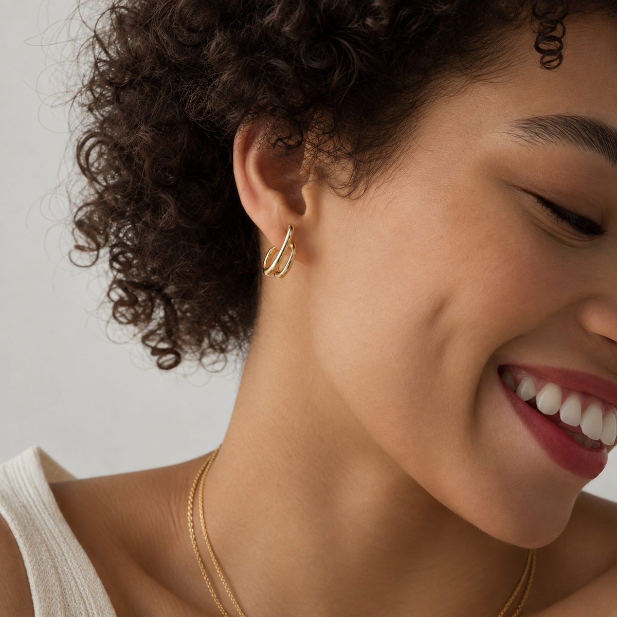 A close-up lifestyle shot of a smiling woman with curly dark hair wearing a delicate gold earring and a thin gold necklace against a light gray background.