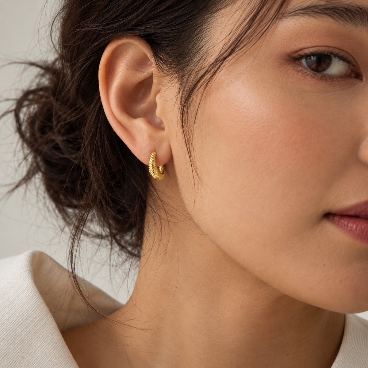 A close-up of a woman's ear wearing a small textured gold huggie hoop earring, with her face and hair partially visible against a soft light background.