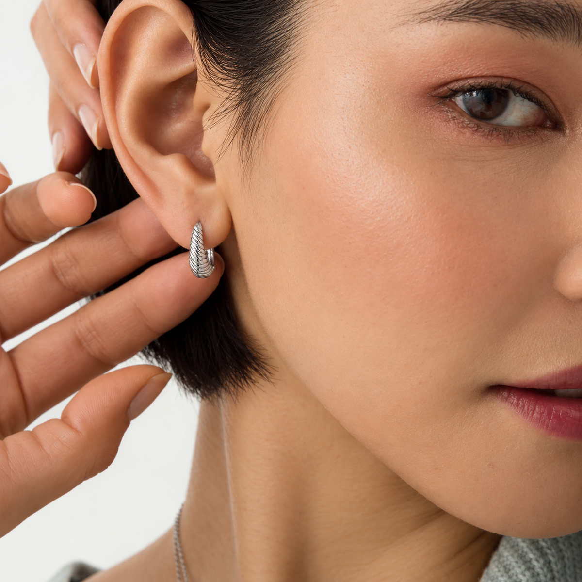 A close-up lifestyle shot of a model wearing a small silver huggie hoop earring with a textured, diamond-like pavé setting. A hand is gently touching the earring to show its scale, against a clean white background.
