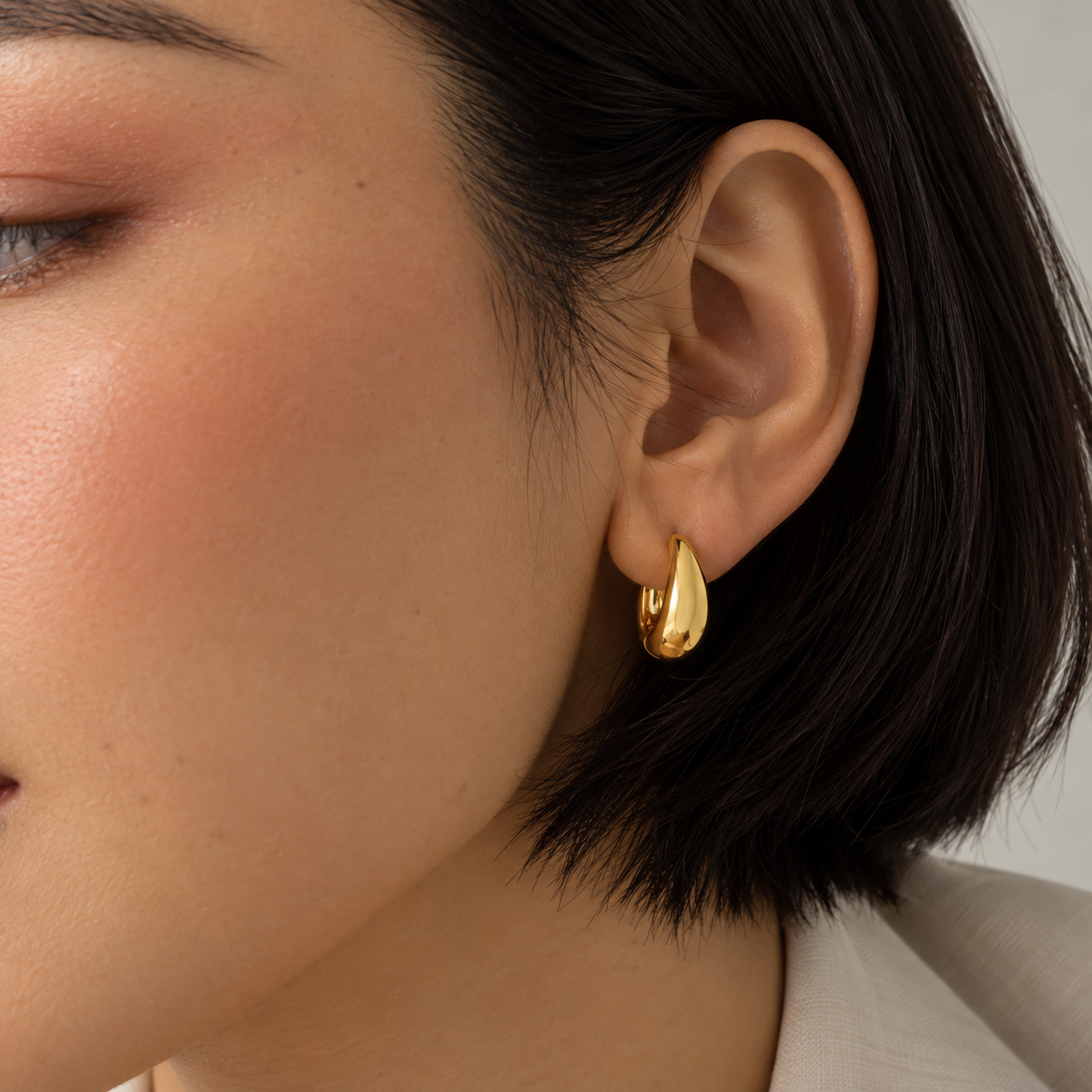 A close-up of a model's ear wearing a small, polished gold huggie hoop earring.