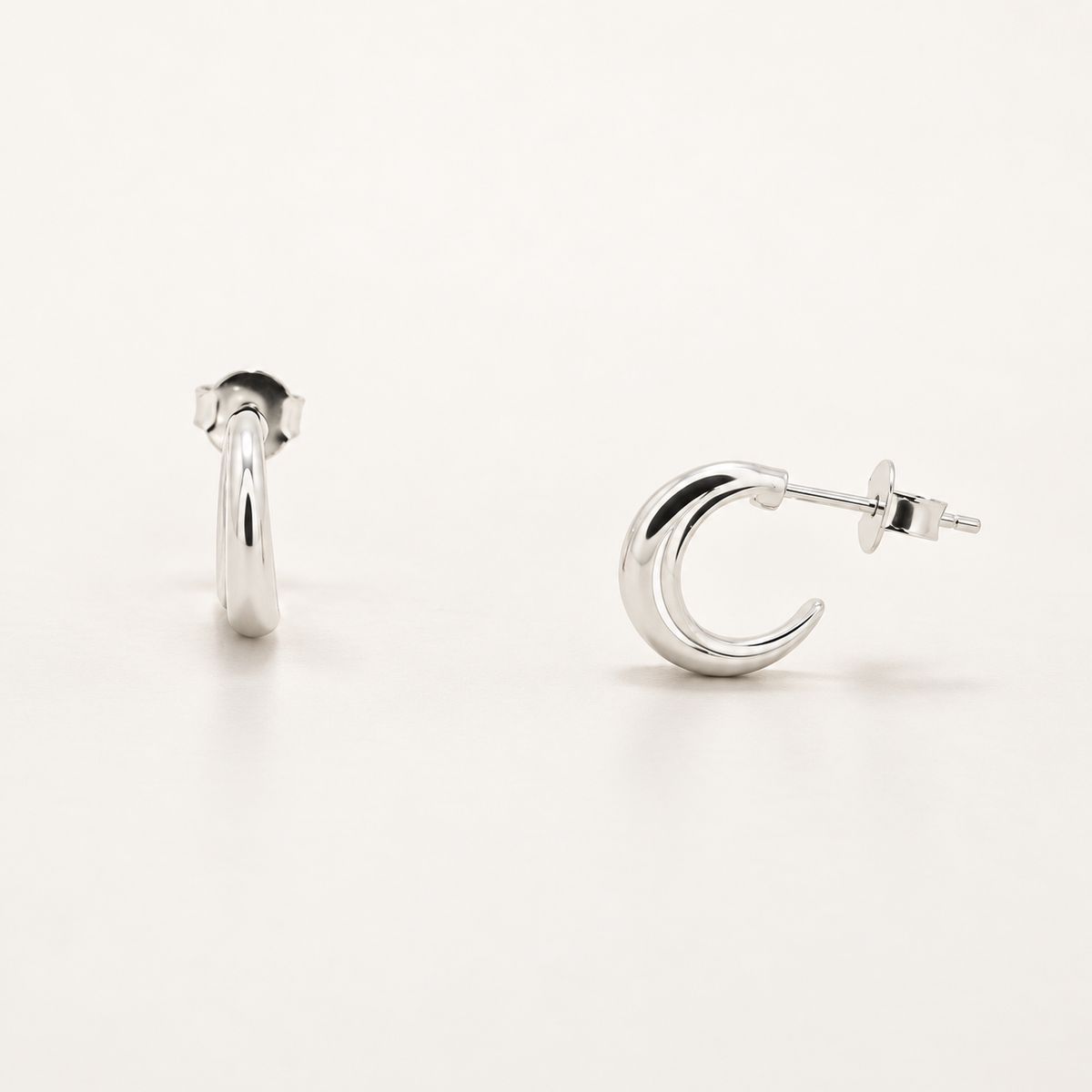 A pair of polished silver hoop earrings shown on a clean white background, with one earring standing upright and the other lying on its side to show the post and backing.