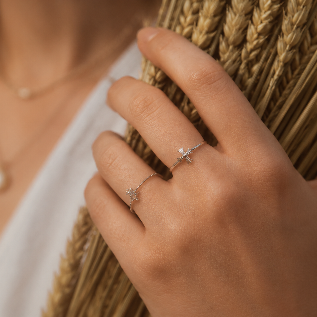 A close-up lifestyle shot of a person's hand resting on a bundle of wheat stalks, wearing two delicate silver rings with star-shaped details.