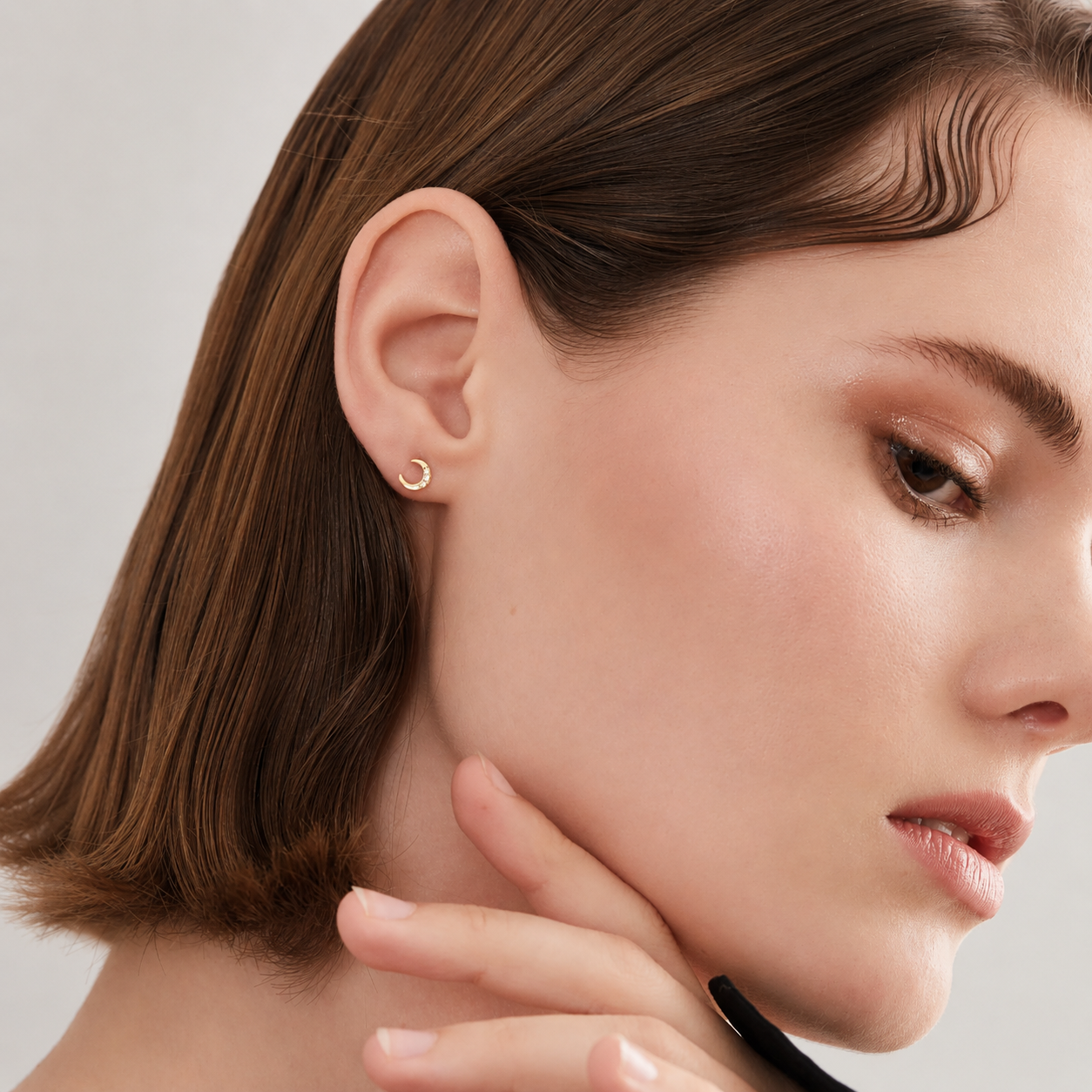 A close-up of a woman's ear wearing a small gold crescent moon stud earring against a soft light gray background.