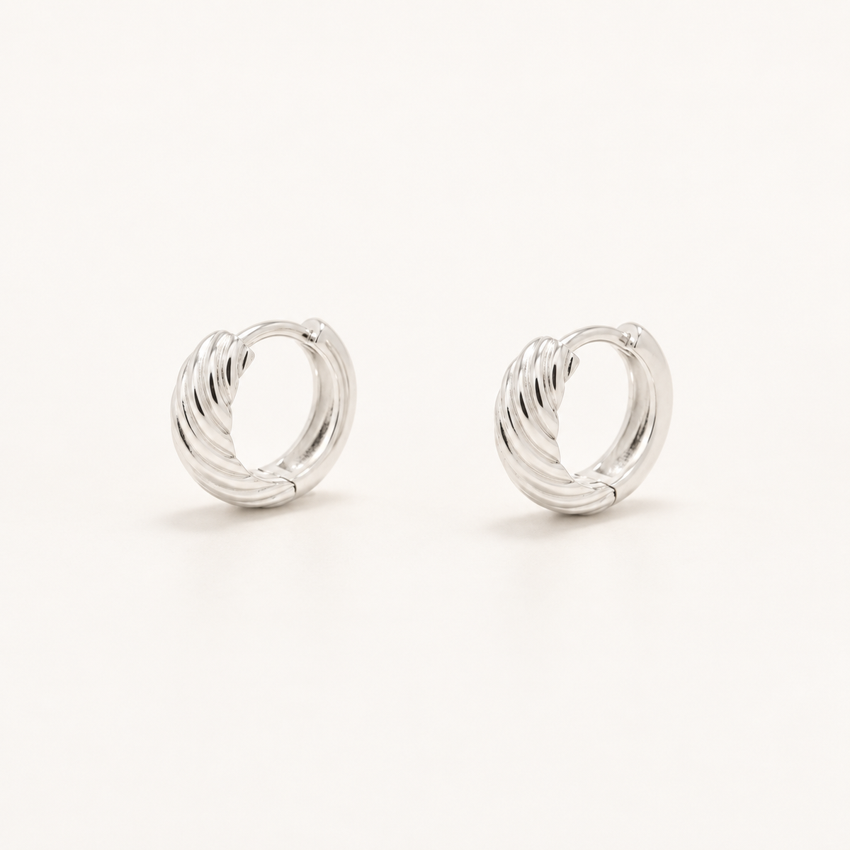 A pair of polished silver huggie hoop earrings with a textured, ribbed spiral design, displayed on a clean off-white background.